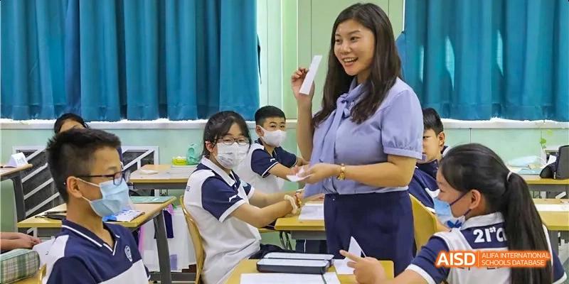 A teacher standing in front of students in a classroom at Beijing 21st Century School, holding a small object and speaking.