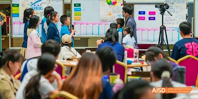 Students and a teacher interacting in a brightly lit classroom at Beijing 21st Century School, with colorful decorations on the wall.