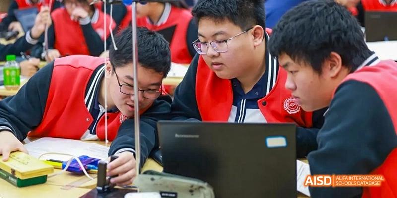 Students in uniform engaged in an activity, looking at a laptop and holding beakers, at Beijing 21st Century School.