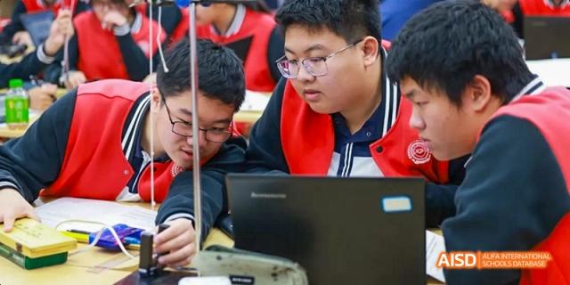 Students in uniform engaged in an activity, looking at a laptop and holding beakers, at Beijing 21st Century School.