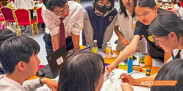 Students gathered around a table, working on a project with various materials and a laptop at Beijing 21st Century School.