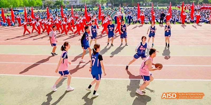 A sports field is filled with students in various uniforms playing a game, with a large group holding red flags in the background at Beijing 101 Middle School.