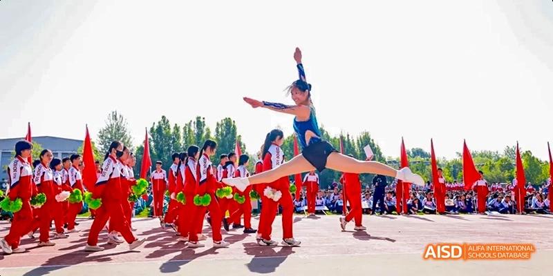 A female athlete in a blue leotard is performing a high kick or jump while other students in red uniforms are sitting in the background at Beijing 101 Middle School.