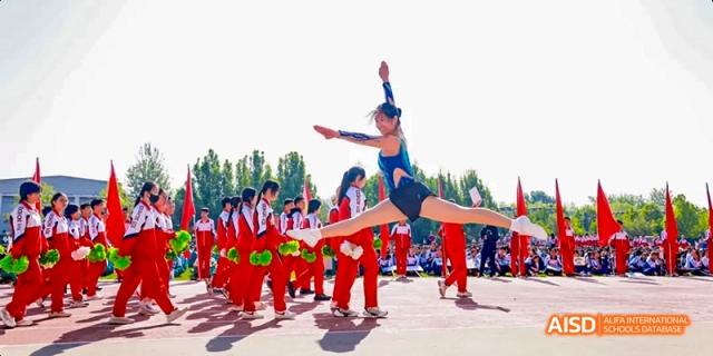 A female athlete in a blue leotard is performing a high kick or jump while other students in red uniforms are sitting in the background at Beijing 101 Middle School.
