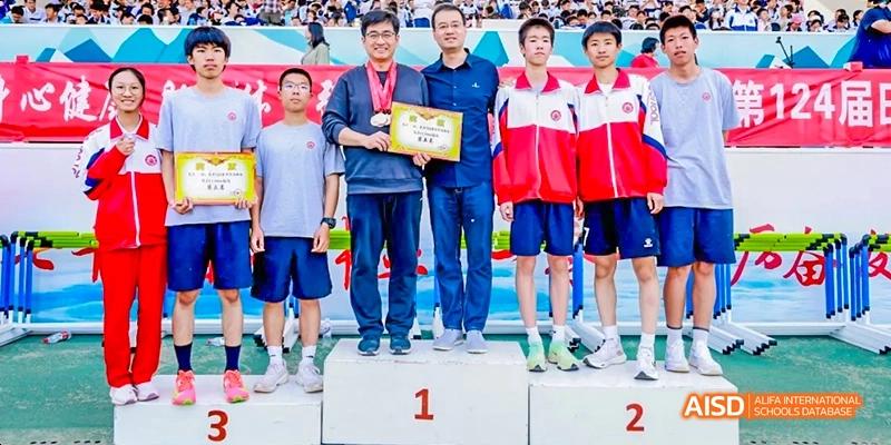Two male students in athletic uniforms are standing on a podium with a teacher and a coach, holding a certificate, at an awards ceremony at Beijing 101 Middle School.