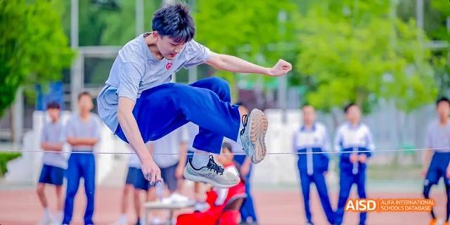 A male student in a blue shirt and white shorts is performing a high kick on a sports field at Beijing 101 Middle School.