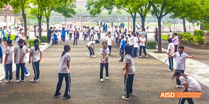 Beijing Bacui Bilingual School A group of students in a schoolyard, some playing a game on a paved area, with other students and trees in the background.