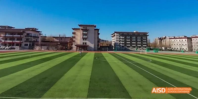 Beijing Bacui Bilingual School A wide view of an empty green athletic field and running track with a school building in the background under a clear sky.