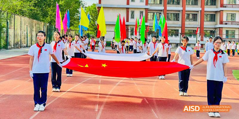 Beijing Bacui Bilingual School A group of students carrying a large Chinese flag and other colorful flags on a running track during an event.