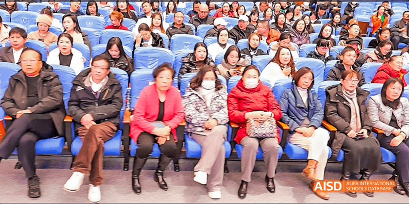 Beijing Bacui Bilingual School An audience of people, including many adults, sits in rows and watches a stage performance.