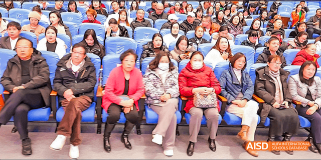 Beijing Bacui Bilingual School An audience of people, including many adults, sits in rows and watches a stage performance.