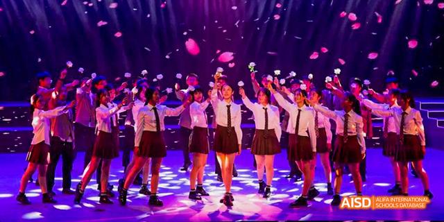 A large group of students from Beijing Haidian Foreign Language Academy in dark skirts and white shirts on a stage, with one of them holding a bouquet of flowers and confetti falling from above.