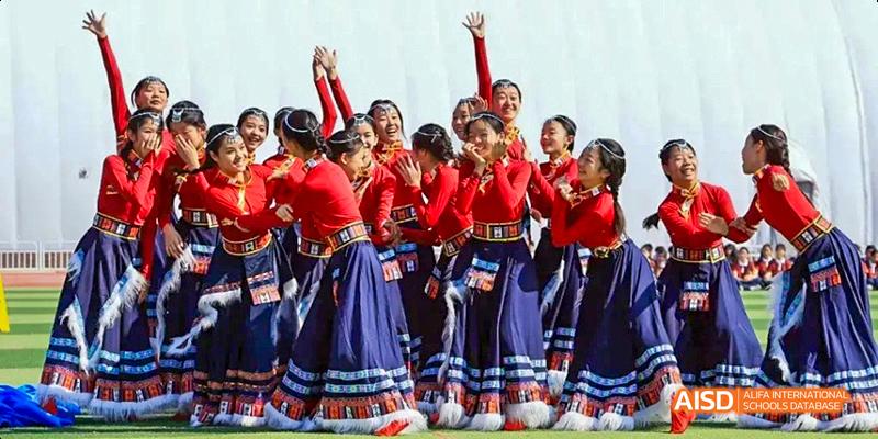 A group of female students from Beijing Haidian Foreign Language Academy performing a traditional dance on a stage, dressed in red tops and blue skirts.