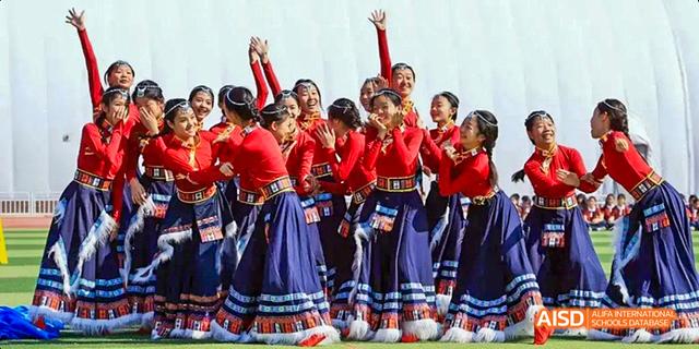 A group of female students from Beijing Haidian Foreign Language Academy performing a traditional dance on a stage, dressed in red tops and blue skirts.