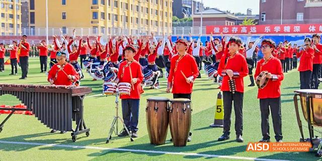 Students from Beijing Haidian Foreign Language Academy dressed in red shirts, playing drums and percussion instruments on a field.