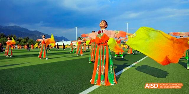 A group of students from Beijing Haidian Foreign Language Academy performing a traditional dance in colorful orange and green costumes with flags.
