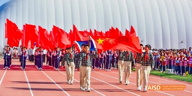 Students from Beijing Haidian Foreign Language Academy in plaid uniforms marching in formation on a track, carrying large red flags.