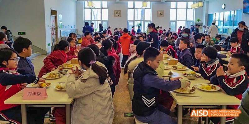 Students sitting at tables in a brightly lit cafeteria, eating and socializing at Beijing Hui Xian School.