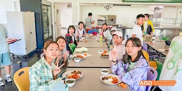 A group of students from Beijing Huijia Private School smile and wave while sitting at a table enjoying a meal together.