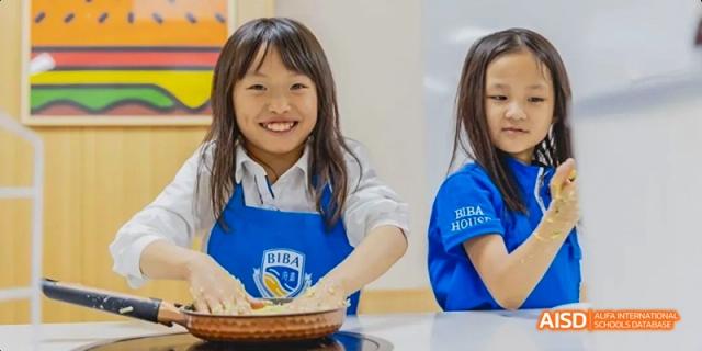 Two young female students, one wearing a BIBA apron, are smiling while preparing food during a cooking class at Beijing International Bilingual Academy.