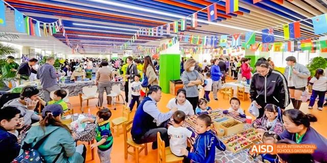 A large, colorful indoor event hall is filled with families and students gathered around food stalls at Beijing International Bilingual Academy.