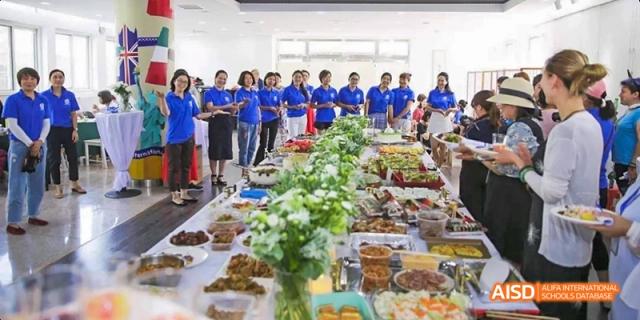 A group of students in blue shirts are posing behind a long buffet table filled with a variety of food at Beijing International Bilingual Academy.