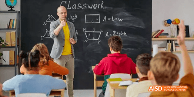 A male teacher from Beilu Academy, standing in front of a blackboard with the word "Classwork" written on it and talking to a student.
