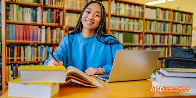 A female student from Beilu Academy, sitting in a library with a laptop and a book, and smiling at the camera.
