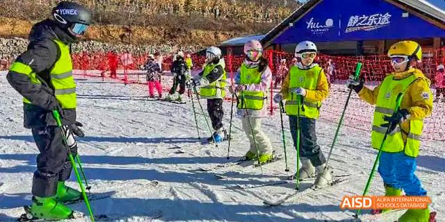 A group of students from Beilu Academy, wearing helmets and bright yellow jackets, learning to ski on a snowy slope.