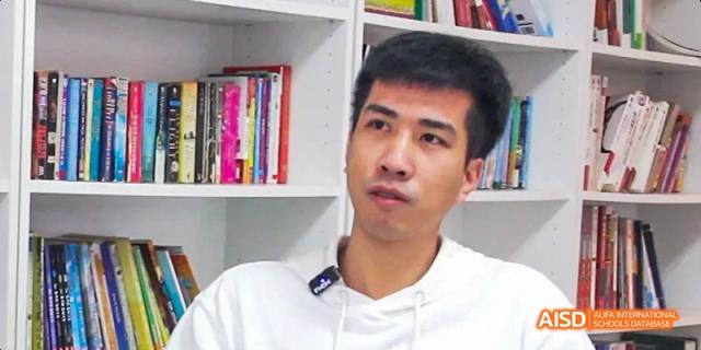 Camford Royal School teacher or staff member speaking in front of a bookshelf.