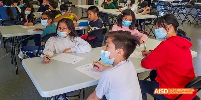 A young person at Dalian American International School with a face mask on, sitting at a desk and writing.