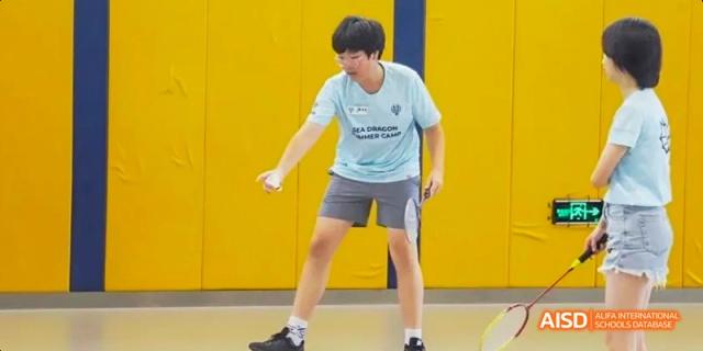 A boy in a blue t-shirt and grey shorts playing table tennis in a gym. He is wearing sneakers and holding a paddle. He's at the Dalian American International School.