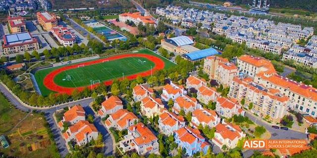 An aerial view of a suburban neighborhood with red-roofed houses and a sports field, including a track and a soccer pitch. This area is associated with the Dalian American International School.