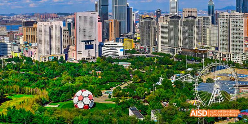 An aerial view of a city park and skyscrapers on a sunny day. Green trees fill the park, and a soccer ball is visible in the grass. This is near the Dalian American International School.