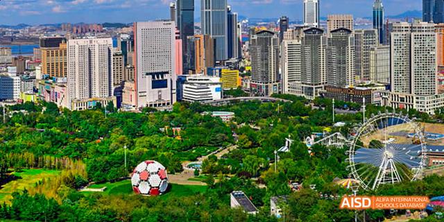 An aerial view of a city park and skyscrapers on a sunny day. Green trees fill the park, and a soccer ball is visible in the grass. This is near the Dalian American International School.
