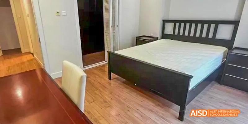 A clean, modern bedroom with wood floors. A bed with a black frame and a simple headboard is against one wall. There's a nightstand on one side. This is a room at the Dalian American International School.