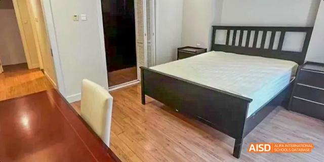 A clean, modern bedroom with wood floors. A bed with a black frame and a simple headboard is against one wall. There's a nightstand on one side. This is a room at the Dalian American International School.