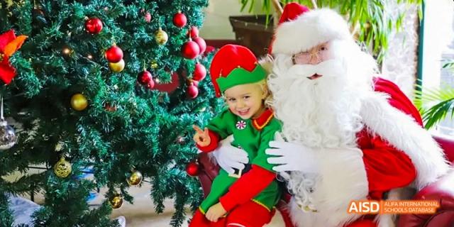 A young child dressed as an elf sitting on Santa's lap next to a decorated Christmas tree. The child is smiling happily. This is a holiday event at the Dalian American International School.