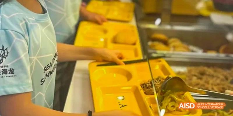 A student holding a yellow plastic tray with multiple compartments. Their hand is in the frame, but the face is out of focus. This is a cafeteria scene at the Dalian American International School.