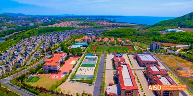 An aerial view of a large coastal community with many buildings, green spaces, and a sports field. The ocean is in the background. This is a location related to the Dalian American International School.