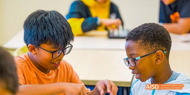 A group of students from diverse backgrounds are sitting at a table. Two are looking at a tablet or book, with a focus on their faces and hands. This is a classroom scene at the Dalian American International School.
