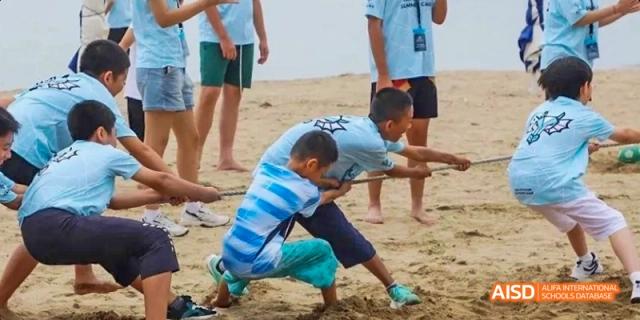 A young person helping another do stretches on a sandy beach at Dalian American International School.
