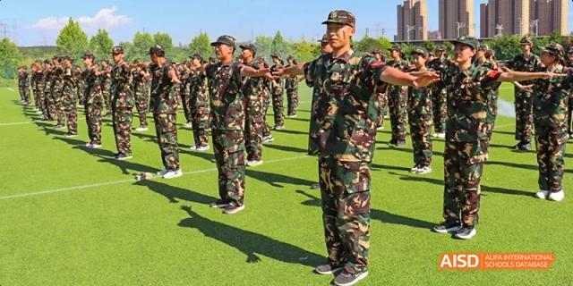 A group of Ealing International School students in camouflage uniforms are doing military-style drills on a field.