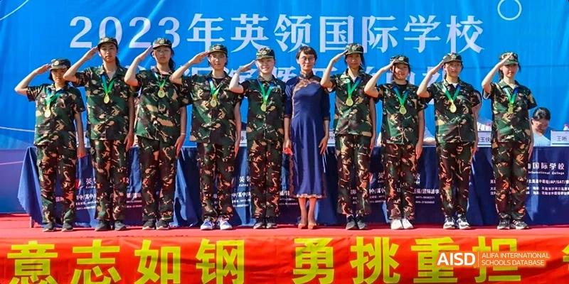 Ealing International School students in camouflage uniforms are standing on a stage with an adult in a blue dress.