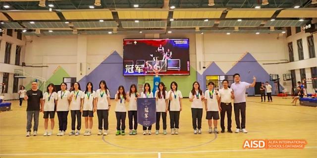 A team of students from HAIDE Bilingual School Dongguan stands in a line with a coach on a basketball court, posing with medals and a championship banner.
