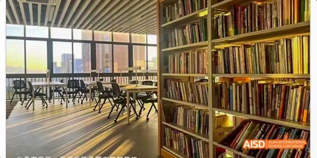 An empty library at HAIDE Bilingual School Dongguan showing a view of the bookshelves, tables, and chairs near a window.