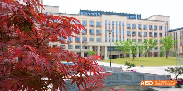 The modern academic building of Hangzhou Foreign Languages School, partially framed by vibrant red maple leaves in the foreground.
