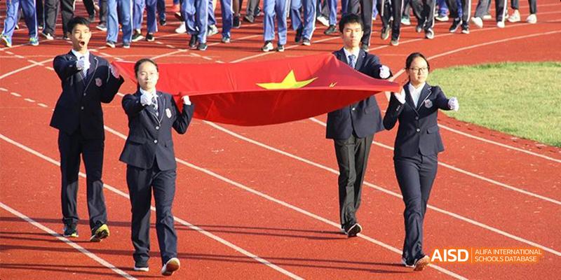 Hangzhou Foreign Languages School students and staff carrying a large Chinese national flag across a red running track, likely during an opening ceremony.