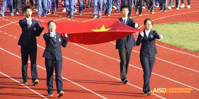 Hangzhou Foreign Languages School students and staff carrying a large Chinese national flag across a red running track, likely during an opening ceremony.