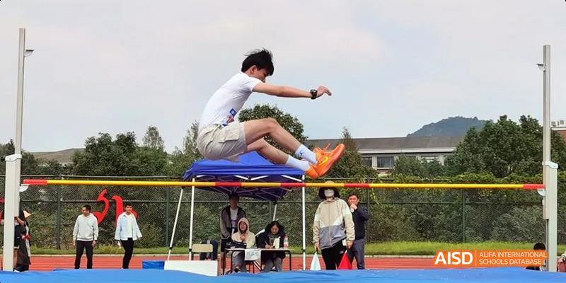 A Hangzhou Foreign Languages School male student in mid-air during a long jump or triple jump competition, with spectators watching in the background.
